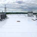 Reflective white membrane roof of a cannabis THCP processing facility with rigid insulation panels, exposed vapor-barrier seam tape, sealed penetrations, stainless HVAC units and ventilation stacks with scrubber housings, and metal parapets under bright overcast light; distant industrial park softly visible.