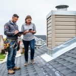 A homeowner and roofer inspecting a newly installed shingle roof near chimney flashing while documenting details with a clipboard on a Canadian residential property.